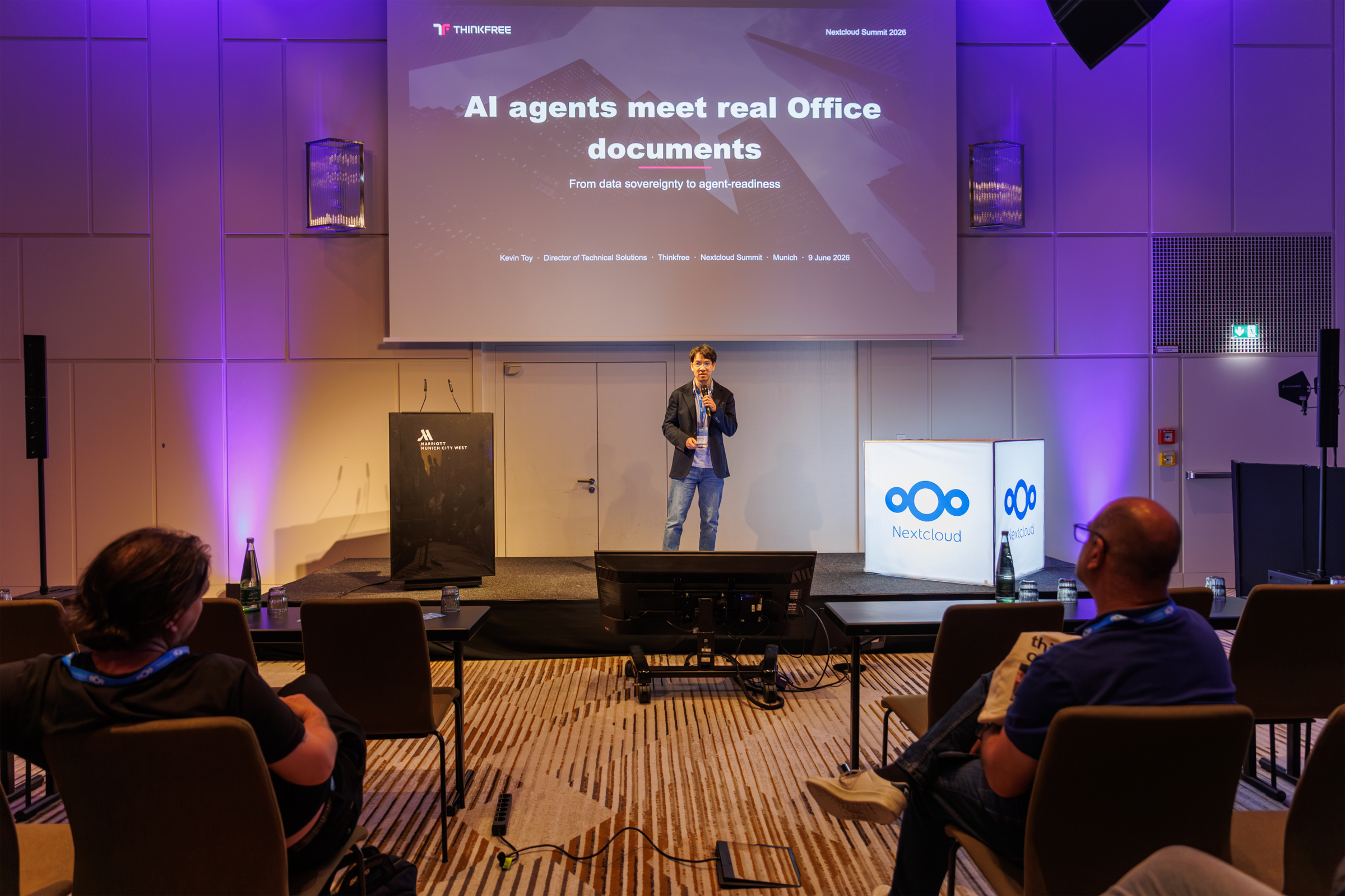 Thinkfree's director of Technical Solutions (Kevin Toy) stands on stage presenting "AI agents meet real Office documents". To his left, a large screen clearly displays a technical schematic titled "Agent-Ready Document Workflows" showing a flow from "AI AGENT" through "THINKFREE AI OFFICE SDK" to "REAL OFFICE DOCUMENTS". An audience listens from the foreground.
