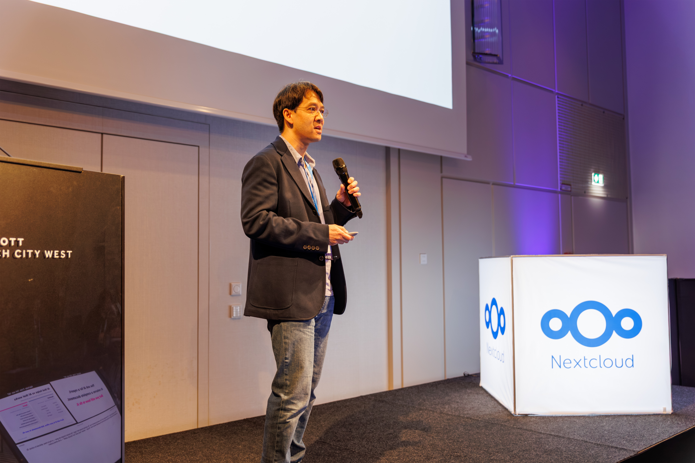 Kevin Toy, Director of Technical Solutions at Thinkfree, delivering a presentation on stage at the Nextcloud Summit 2026 in Munich, standing next to a glowing Nextcloud logo cube.